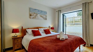 Une chambre avec un grand lit, deux tables de chevet, un tableau au mur et une vue sur l’extérieur par une porte ouverte.