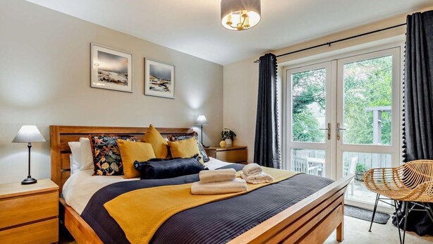 A bedroom with a wooden bed, a nightstand, a lamp, a window with curtains, and a view of greenery outside. - 3 Bed in Morpeth (oc-91148) (Rothbury)