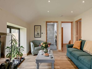 A living room with a sofa, a table with a vase of flowers, and a kitchen area with a refrigerator. - 2 Bed in Norwich (oc-91298) (Corpusty)