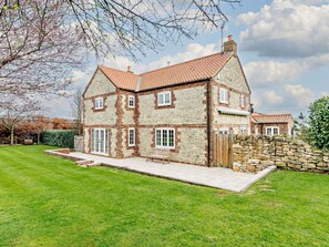 A two-story stone house with a tiled roof, a stone wall, and a grassy lawn.