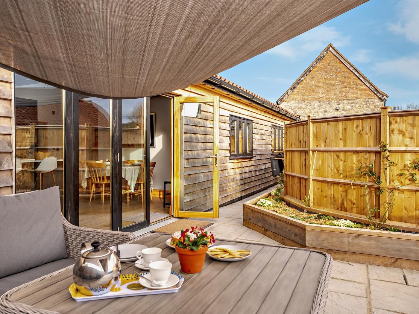 A cozy outdoor seating area with a table set for tea, a wooden fence, and a building with large windows.