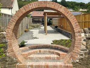 A brick archway leading to a patio area with a barbecue grill, a wooden fence, and a house in the background. - 2 Bed in Axbridge (oc-91358) (Compton Bishop)
