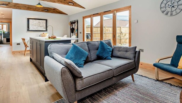 A modern living room with a grey sofa, wooden ceiling beams, a kitchen area, and a large window. - 2 Bed in Axbridge (oc-91358) (Compton Bishop)