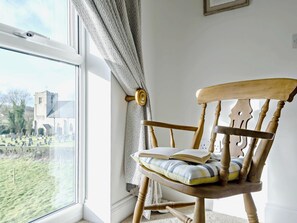 Una mecedora de madera con cojín y una cortina estampada, ubicada cerca de una ventana con vista a una iglesia y un cementerio.