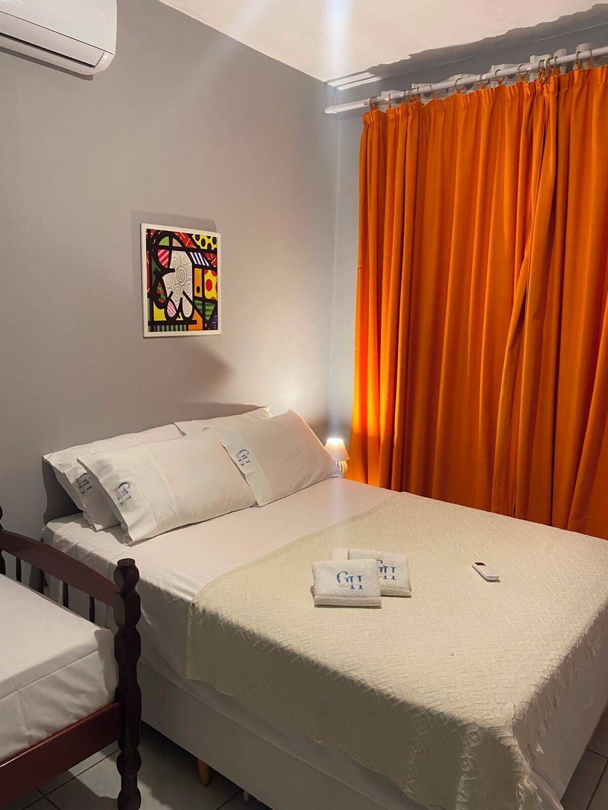 Quarto de hotel com cama, mesa de cabeceira, ar condicionado fixado na parede, um quadro e cortinas laranja.