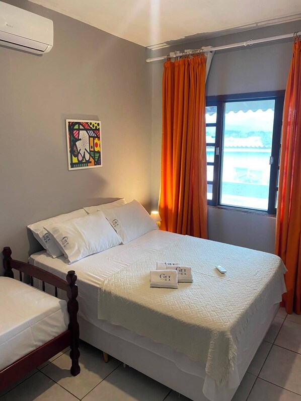 A hotel room with a bed, a window with curtains, a painting on the wall, and an air conditioning unit.