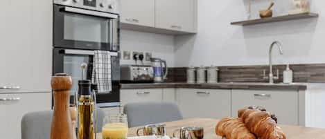 Une cuisine moderne avec une table à manger dressée pour le petit-déjeuner, comprenant des croissants, des tomates et un verre de jus d’orange.