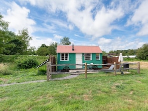 A small, single-story building with a red roof and green walls, situated in a grassy area with a wooden fence and a metal gate. - 1 Bed in Brighton (oc-90450) (Poynings)