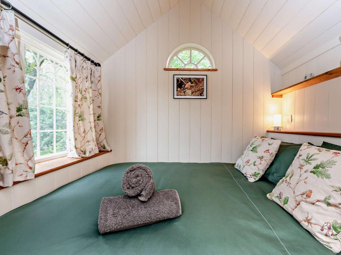 A bedroom with a bed, a window with curtains, a picture on the wall, and a bedside table with a lamp.
