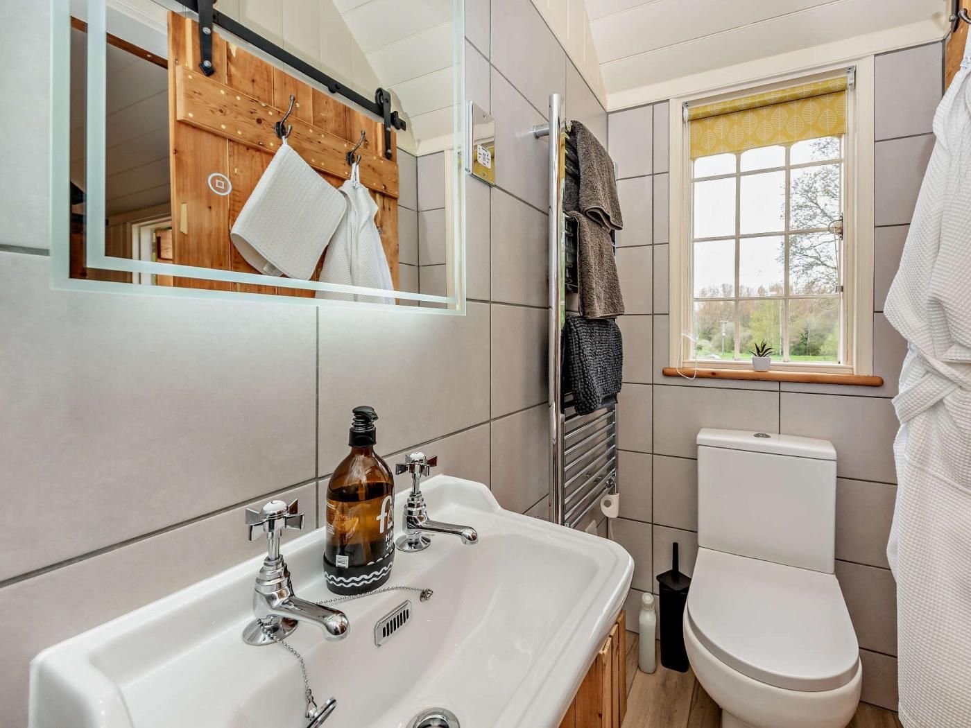 Un baño con lavamanos blanco, un espejo, toallero de madera, inodoro y ventana con vistas a árboles.