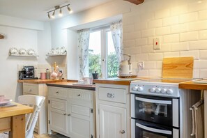 A kitchen with white cabinetry, a stove, a kettle, and a window with floral curtains. - 2 Bed in Exeter (oc-90455) (Tedburn Saint Mary)