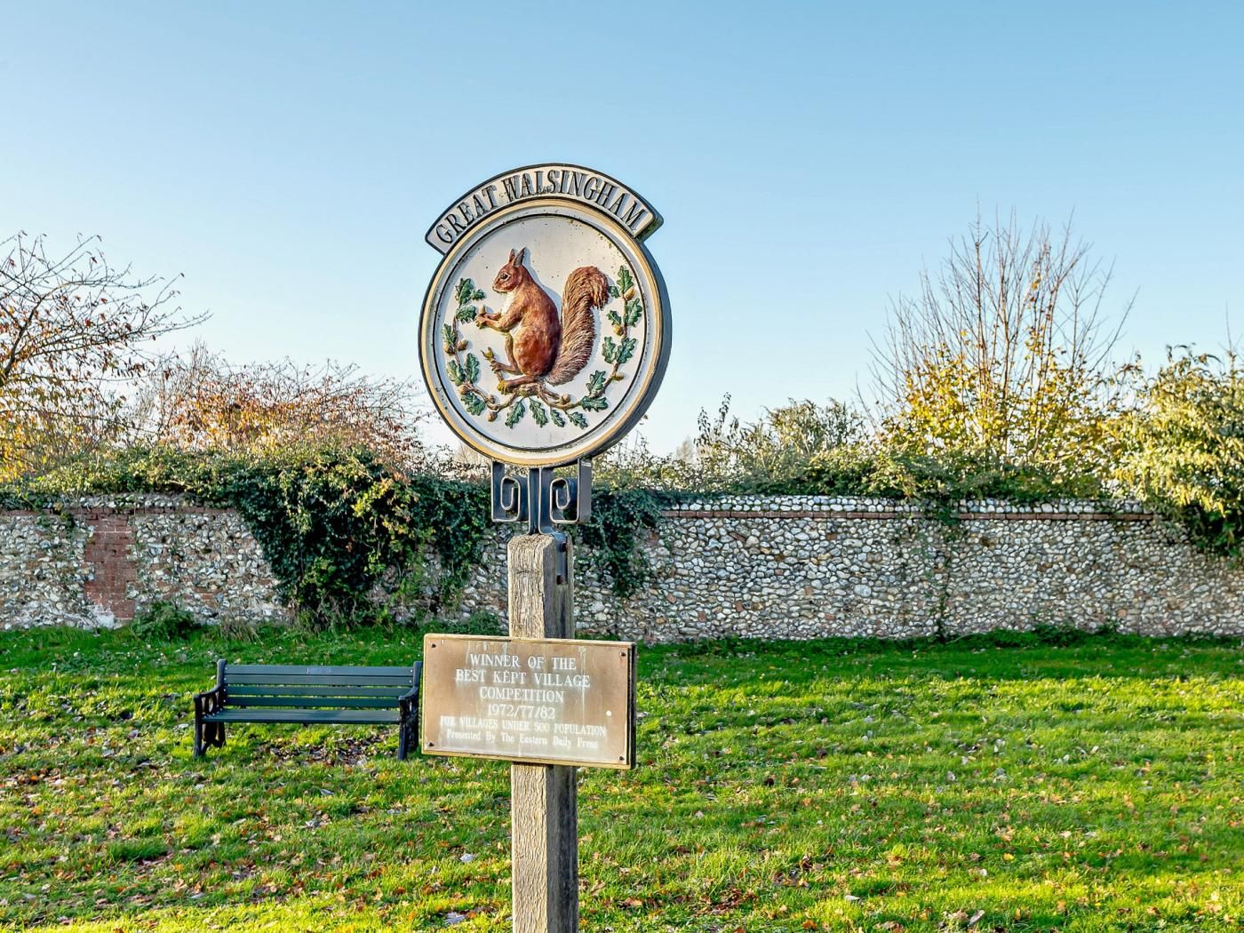 A village sign with a squirrel emblem, a plaque for a village competition winner, and a bench in a grassy area.