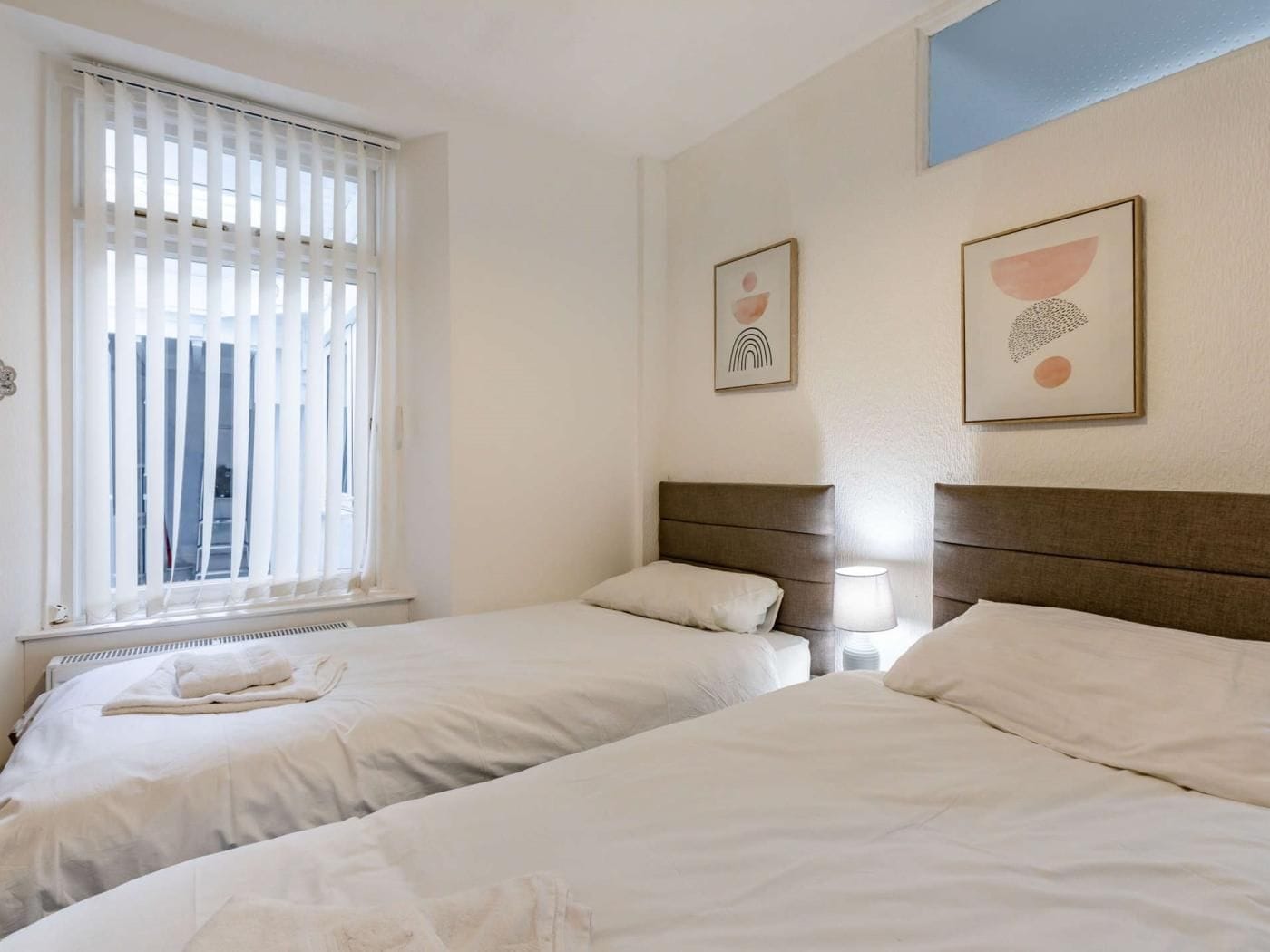 Une chambre d’hôtel avec deux lits, une fenêtre avec des stores, un radiateur et deux œuvres d’art accrochées au mur.