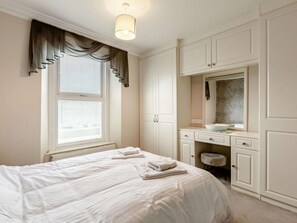 Quarto com cama, janela com cortinas, radiador, penteadeira com espelho e toalha sobre a cama.