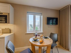 A dining area with a round table, chairs, a television on the wall, a window with a view, and kitchen appliances. - 1 Bed in Ambleside (oc-90637) (Ambleside)