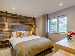 A bedroom with a wooden headboard, a bed with a yellow and white checkered bedspread, bedside lamps, a nightstand, a wooden desk, and a window with a view of trees. - 1 Bed in Ambleside (oc-90637) (Ambleside)