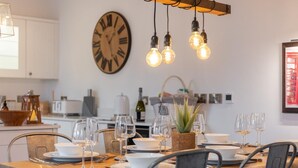 A dining area with a wooden table set for four, surrounded by chairs. A large wall clock and hanging light bulbs are visible. - 3 Bed in Hawes (oc-90749) (Hawes)