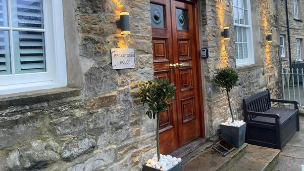 A stone building with a wooden double door, a sign reading 'BECKSTONES HOUSE,' and a bench outside. - 3 Bed in Hawes (oc-90749) (Hawes)