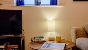 Una habitación con televisión, una mesa pequeña con libros y una lámpara, una ventana con cortinas y un sofá.