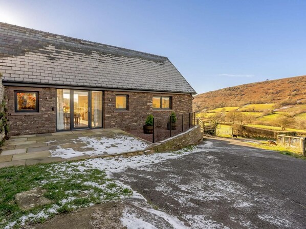 A stone house with a sloped roof, large glass doors, and a paved driveway.