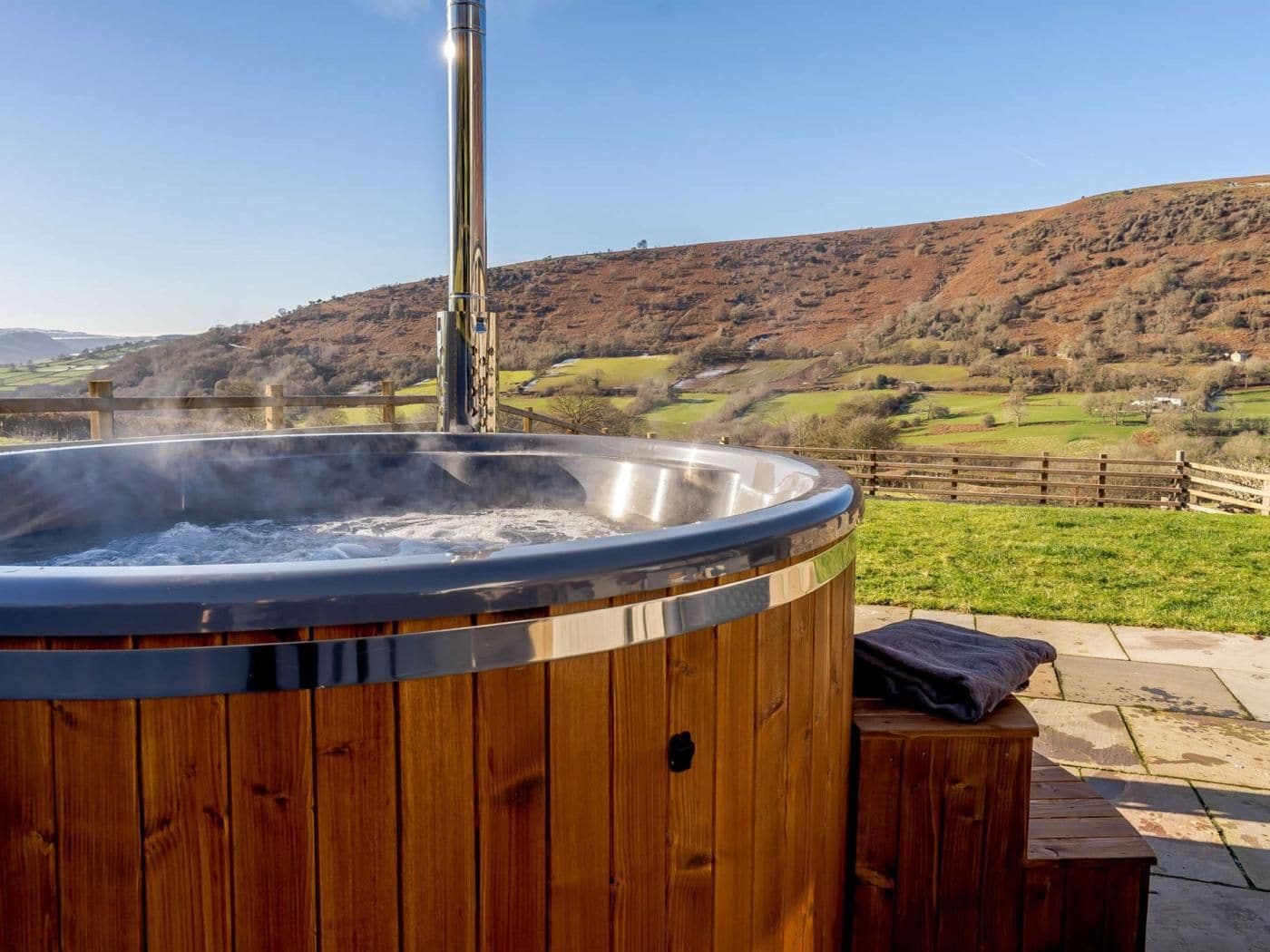 A hot tub with a wooden exterior, surrounded by a wooden deck and a scenic hillside view.