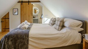 Een slaapkamer met een schuine kap, een bed met witte lakens, geruite kussens, een gebreide deken en een houten nachtkastje met een beker.