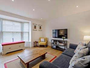 A living room with a sofa, a coffee table, a TV, a bookshelf, and a window with blinds. - 3 Bed in Whitstable (oc-91063) (Whitstable)