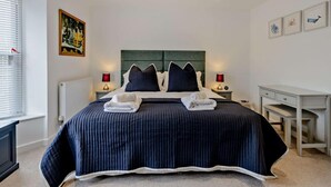 A neatly made bed with a dark blue bedspread, two bedside lamps, a framed artwork above the bed, and a small desk with drawers to the right. - 3 Bed in Whitstable (oc-91063) (Whitstable)