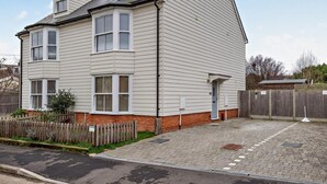 A two-story house with a sloped roof, a brick foundation, and a wooden fence. - 3 Bed in Whitstable (oc-91063) (Whitstable)