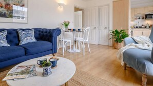 Una sala de estar con un sofá azul, una mesa de centro con revistas, y un comedor con sillas blancas y una mesa.