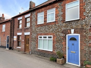 A traditional brick house with a stone facade, a blue door, and a small garden area with potted plants. - 2 Bed in Melton Constable (oc-91371) (Briston)