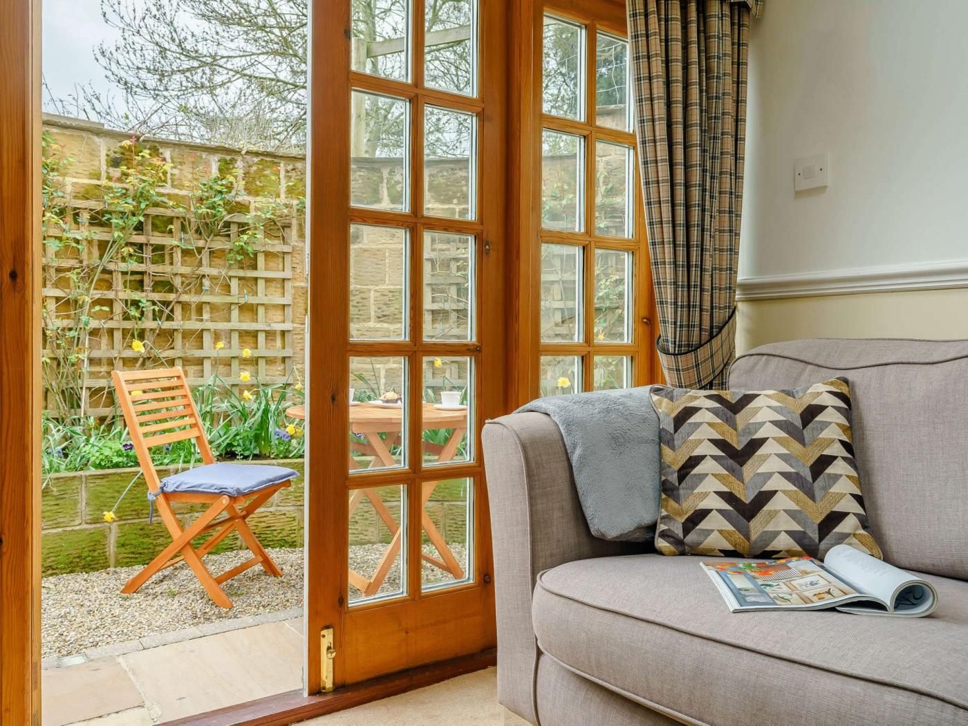 A room with a wooden door leading to a garden, a grey sofa with patterned cushions, and a magazine on the sofa.