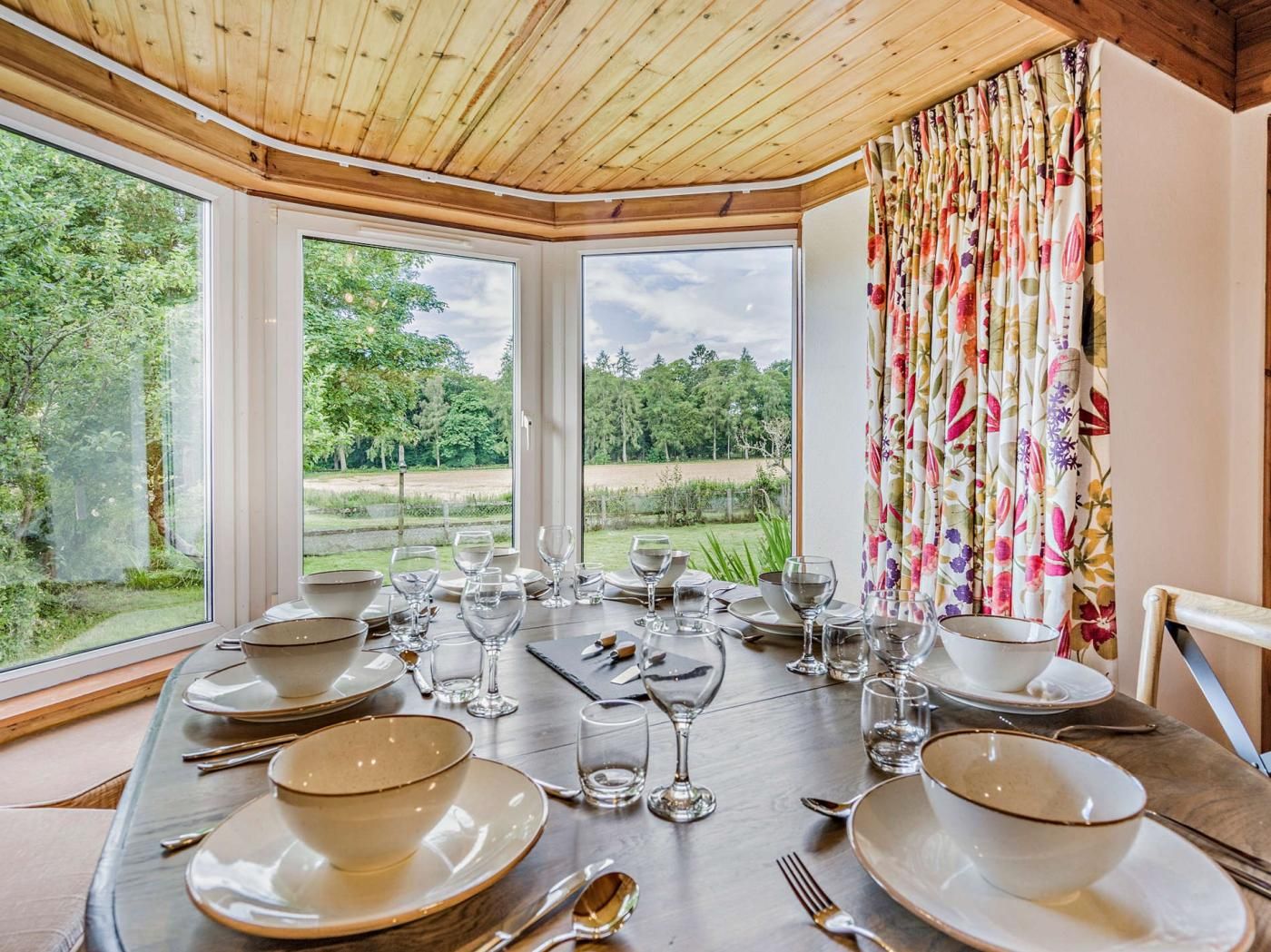 Une table à manger dressée pour six personnes, avec de la vaisselle et des verres, de grandes fenêtres donnant sur un paysage verdoyant et un plafond en bois.