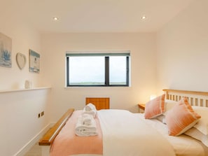 A bedroom with a bed, pillows, a bedside table, a window, and wall art. - 4 Bed in Bude (oc-90539) (Darley)