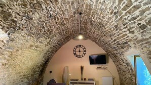 An attic room with a stone ceiling, a clock, a flat-screen TV, a sofa, a dining table with chairs, and a window. - Old stone sheepfold: charm and authenticity in the heart of nature. Dog, okay. (Saint-Beauzély)