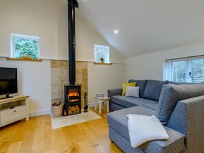 Ein modernes Wohnzimmer mit Kamin aus Stein, einem grauen Sofa, einem Fernseher und einem Fenster mit Blick auf Grünflächen.