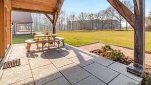 Ett takförsett utomhusområde med ett picknickbord och bänkar, en gräsyta och utsikt över en byggnad i bakgrunden.