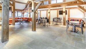 Ett rymligt interiört utrymme med synliga träbjälkar, stengolv och en blandning av olika sittgrupper, inklusive en soffa och bord.