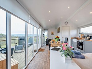 A spacious room with a large sliding glass door leading to an outdoor area, a dining table with chairs, a kitchen area, and a sofa with a view.