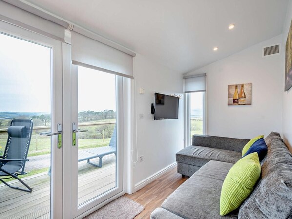 A modern living room with a grey sofa, a television mounted on the wall, and a sliding glass door leading to an outdoor area with a chair and a table.