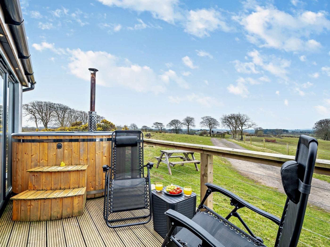A wooden deck with a hot tub, two chairs, and a picnic table with fruit and juice.
