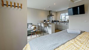 A modern bedroom with a large bed, a TV mounted on the wall, and a kitchenette in the background. - 1 Bed in Barnstaple (oc-91387) (Prixford)