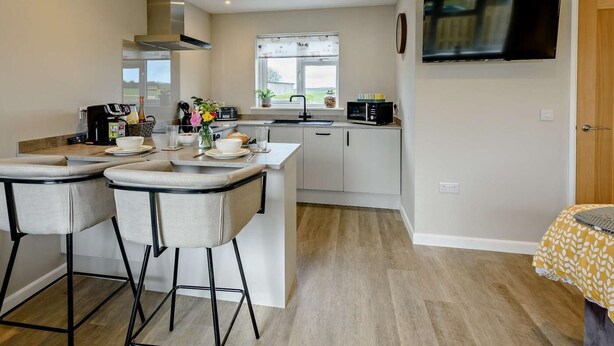 A modern kitchen with a dining area, a TV mounted on the wall, and a window providing natural light. - 1 Bed in Barnstaple (oc-91387) (Prixford)