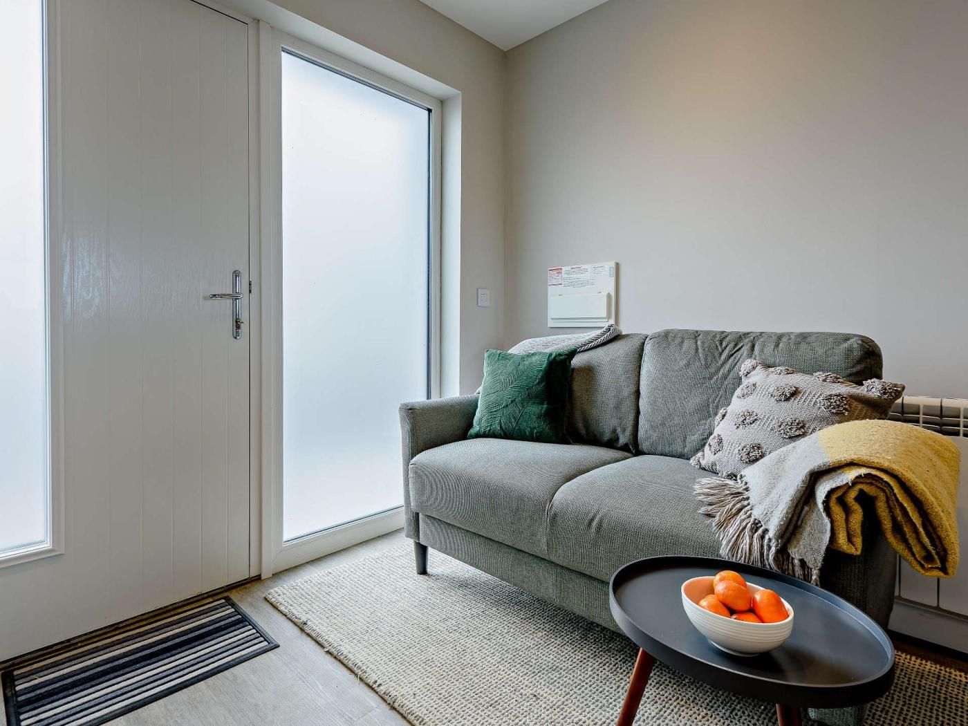 Ein Wohnzimmer mit einem grauen Sofa, einem kleinen runden Tisch mit einer Schale Orangen, einer Heizung und einer Tür mit Glasfenster.