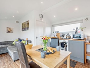 A modern kitchen with a microwave, stove, and sink. There is a dining table with a vase of flowers and a croissant. A gray sofa is in the living area.