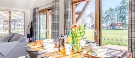 Ein Essbereich mit Tisch, gedeckt für das Frühstück, einer Couch und Blick auf ein grasbewachsenes Feld durch große Fenster.