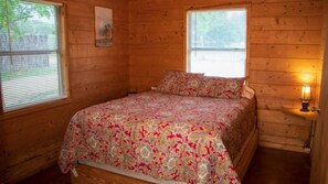 A wooden cabin bedroom with a bed, two windows, and a nightstand with a lamp. - Riverfront Cabins  and  Vacation Homes (Rio Frio)