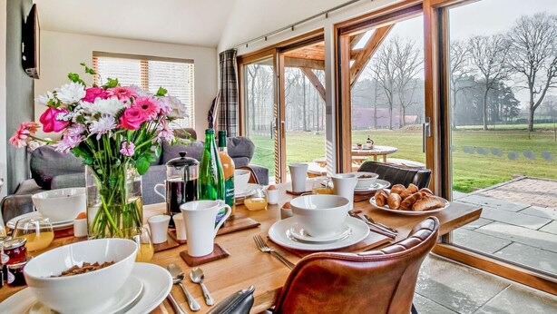 A dining area with a table set for breakfast, a vase of flowers, and a view of the outdoors. - 3 Bed in King's Lynn (oc-91286) (North Runcton)