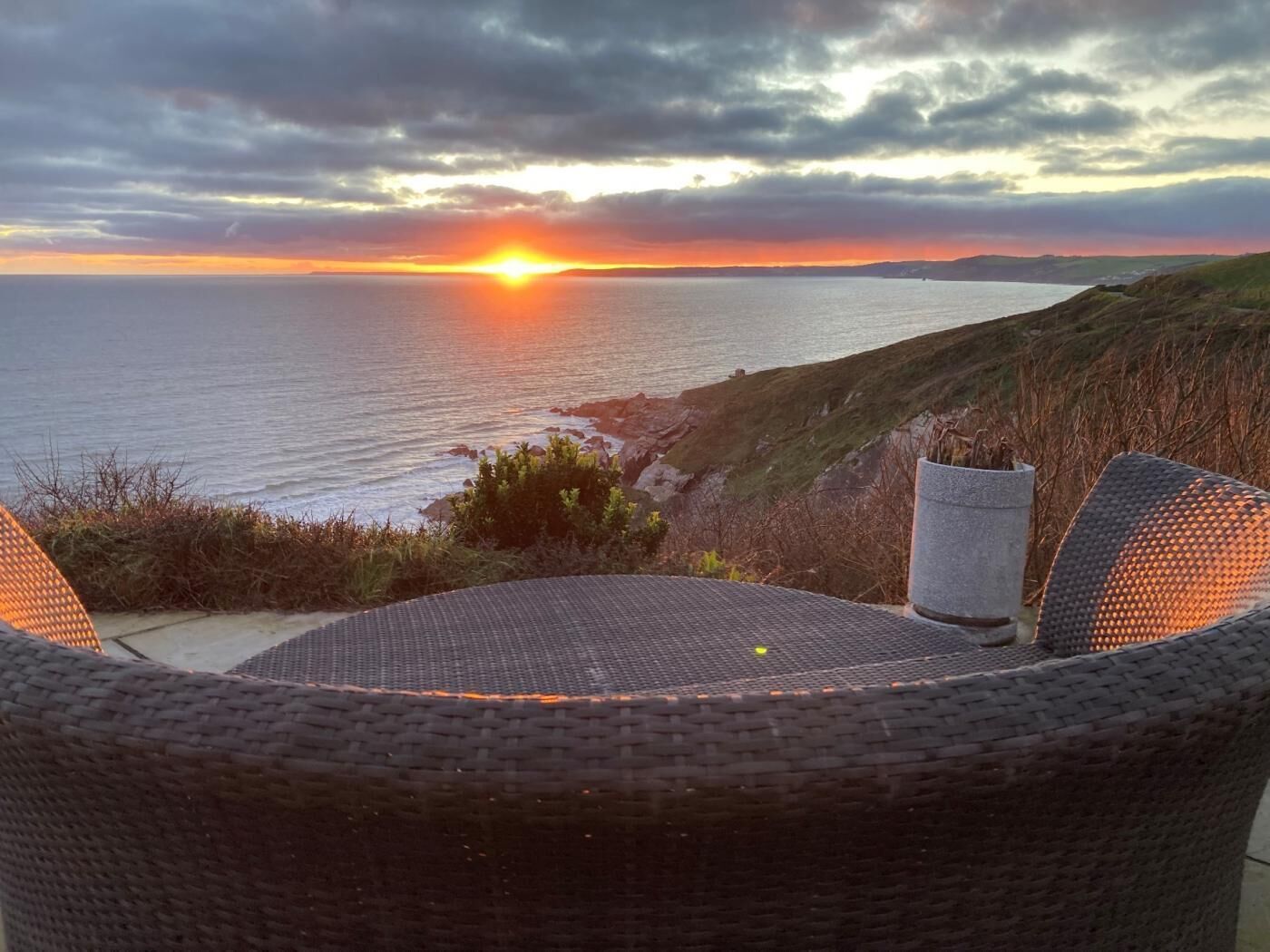 Blick auf das Meer bei Sonnenuntergang, ein Korbstuhl und eine Steinstruktur.