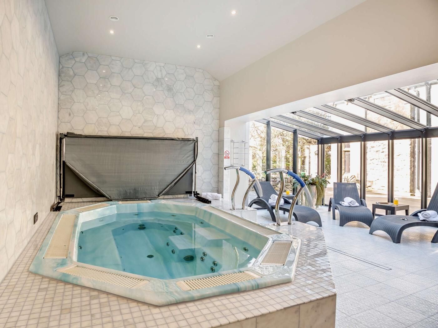 A hot tub with a hexagonal tiled wall, a covered seating area with lounge chairs, and a glass-enclosed patio.
